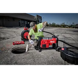 A worker in a green shirt and cap operates a 200’ Stiff Modular Pipeline Inspection Reel by Milwaukee in an outdoor setting. The reel is being used to inspect an underground pipeline, with various tools and equipment visible nearby. The area is a paved surface near a building.