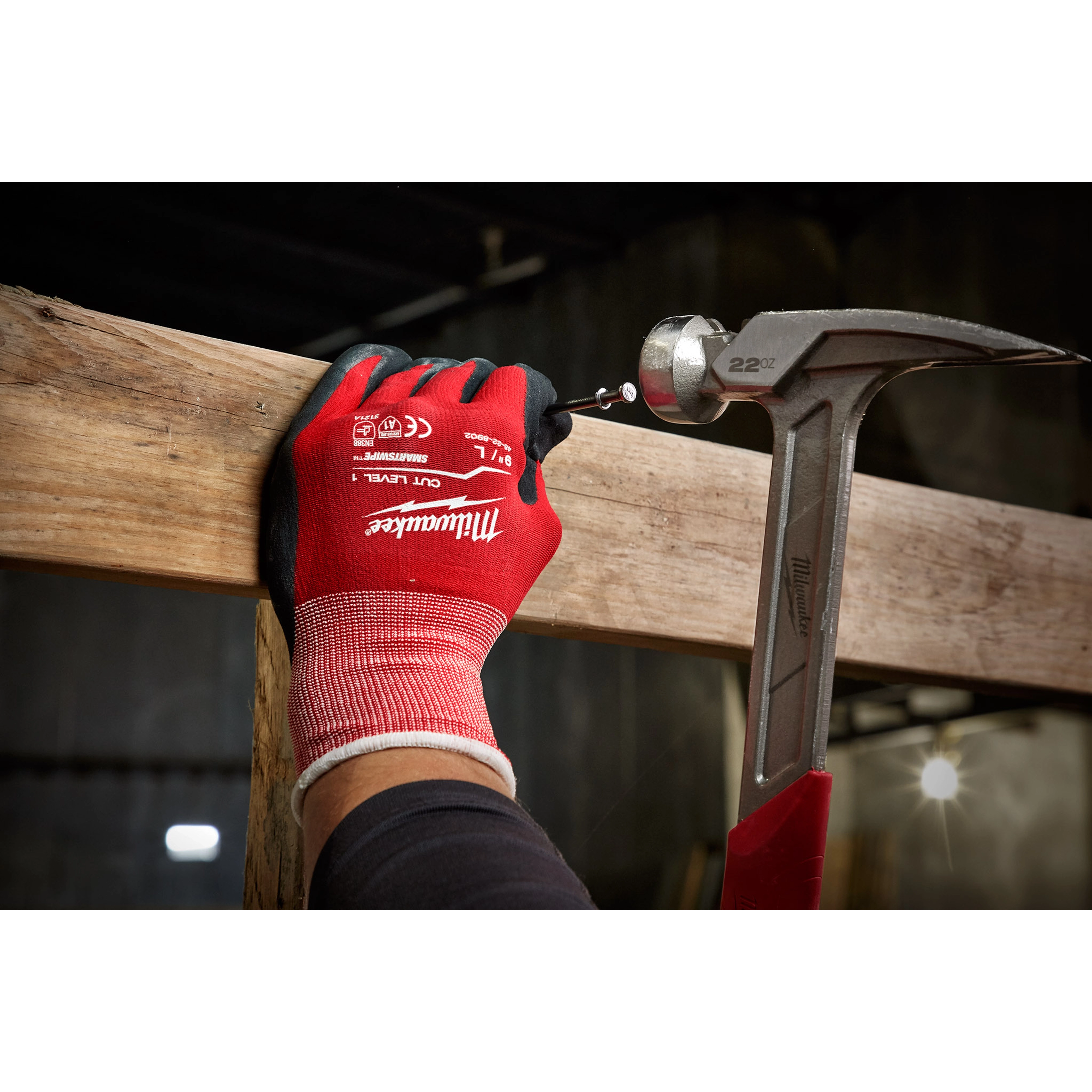 A person wearing red and black Cut%201%20Dipped%20Gloves is holding a wooden plank while using a hammer to drive a screw into it. The scene appears to be in a dimly lit, industrial environment.