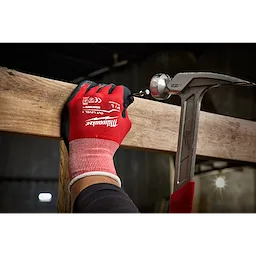 A person wearing red and black Cut%201%20Dipped%20Gloves is holding a wooden plank while using a hammer to drive a screw into it. The scene appears to be in a dimly lit, industrial environment.
