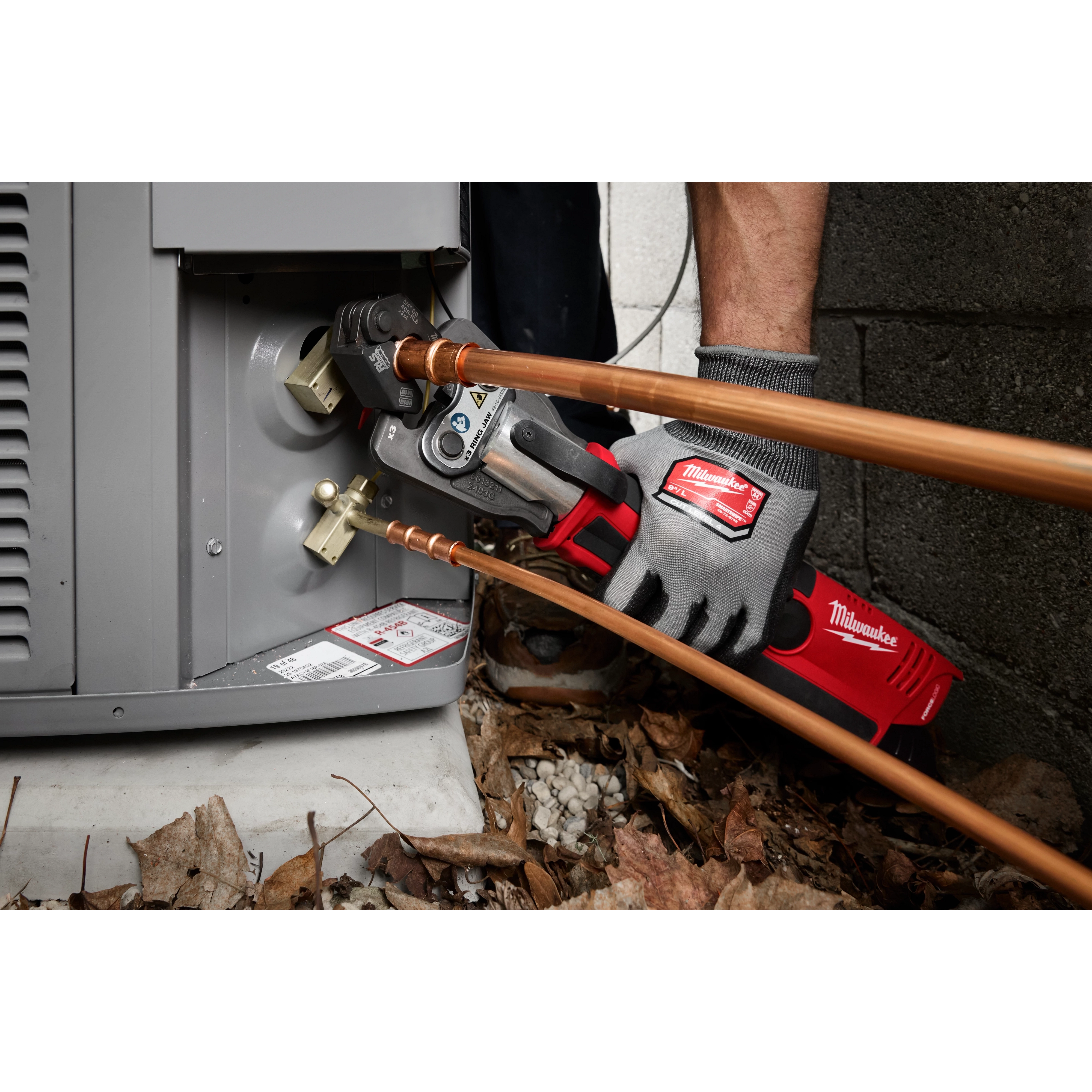 A technician uses a Milwaukee tool equipped with a 3/4" RLS® Pivoting Press Ring to connect copper pipes to an outdoor HVAC unit. The product aids in pressing fittings for a secure connection. The work area is surrounded by dead leaves and gravel.