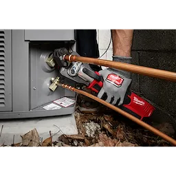 A technician uses a Milwaukee tool equipped with a 3/4" RLS® Pivoting Press Ring to connect copper pipes to an outdoor HVAC unit. The product aids in pressing fittings for a secure connection. The work area is surrounded by dead leaves and gravel.
