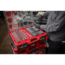A person in work clothes uses a tool from a 98pc 3/8" 6-Pt and 12-Pt Metric & SAE Ratchet & Socket Set with PACKOUT™ Low Profile Organizer. The red and black organizer with multiple compartments is open, displaying various sockets and tools.
