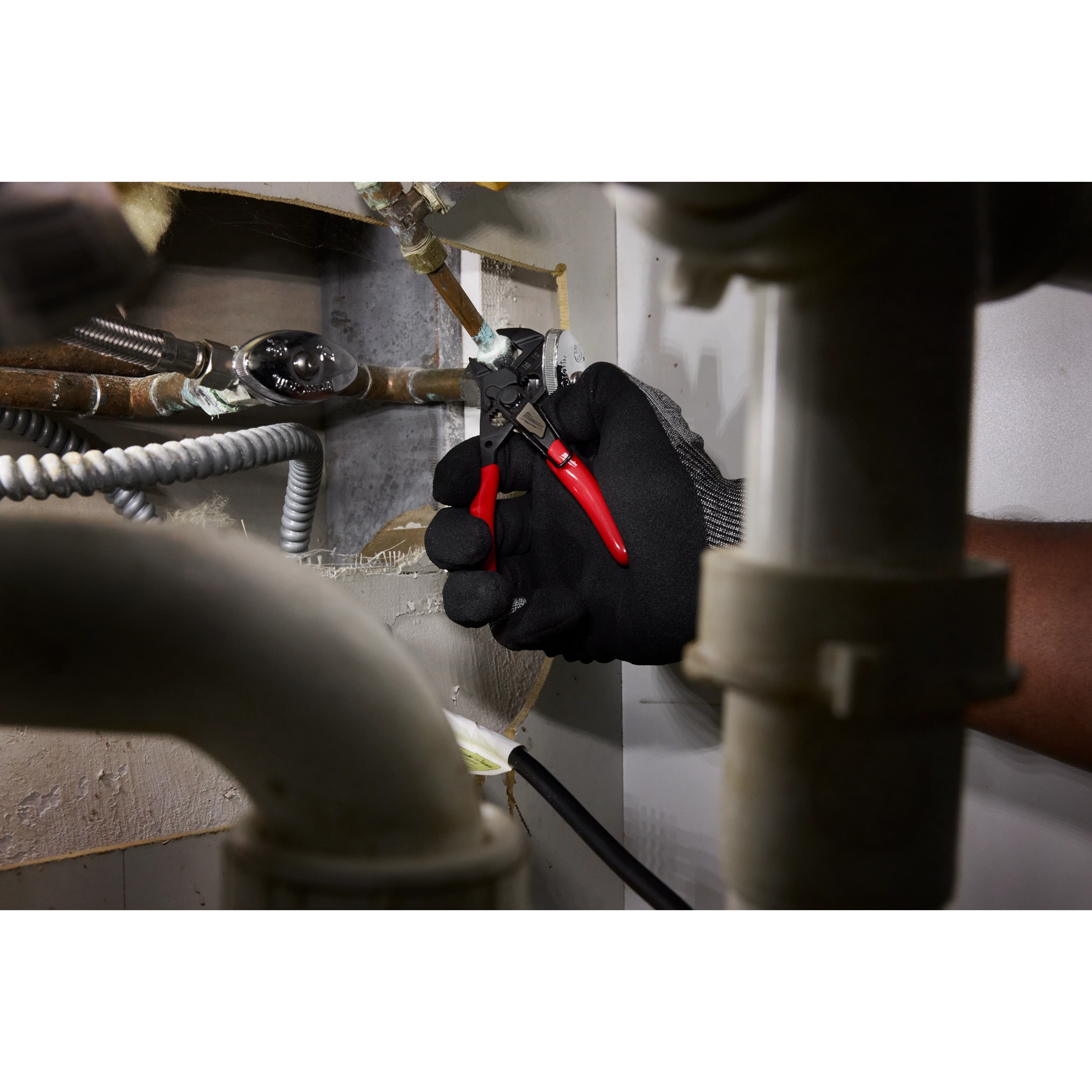 A hand wearing a black glove uses the 5" Pliers Wrench W/ Belt Clip to tighten a metal pipe connection in a tight space, surrounded by various plumbing and electrical components.