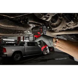A gloved hand uses the M12 FUEL™ 3/8" Subcompact Impact Wrench w/ Friction Ring to work on a vehicle's undercarriage in a garage setting. The compact, red and black tool shows its efficient design for tight spaces.