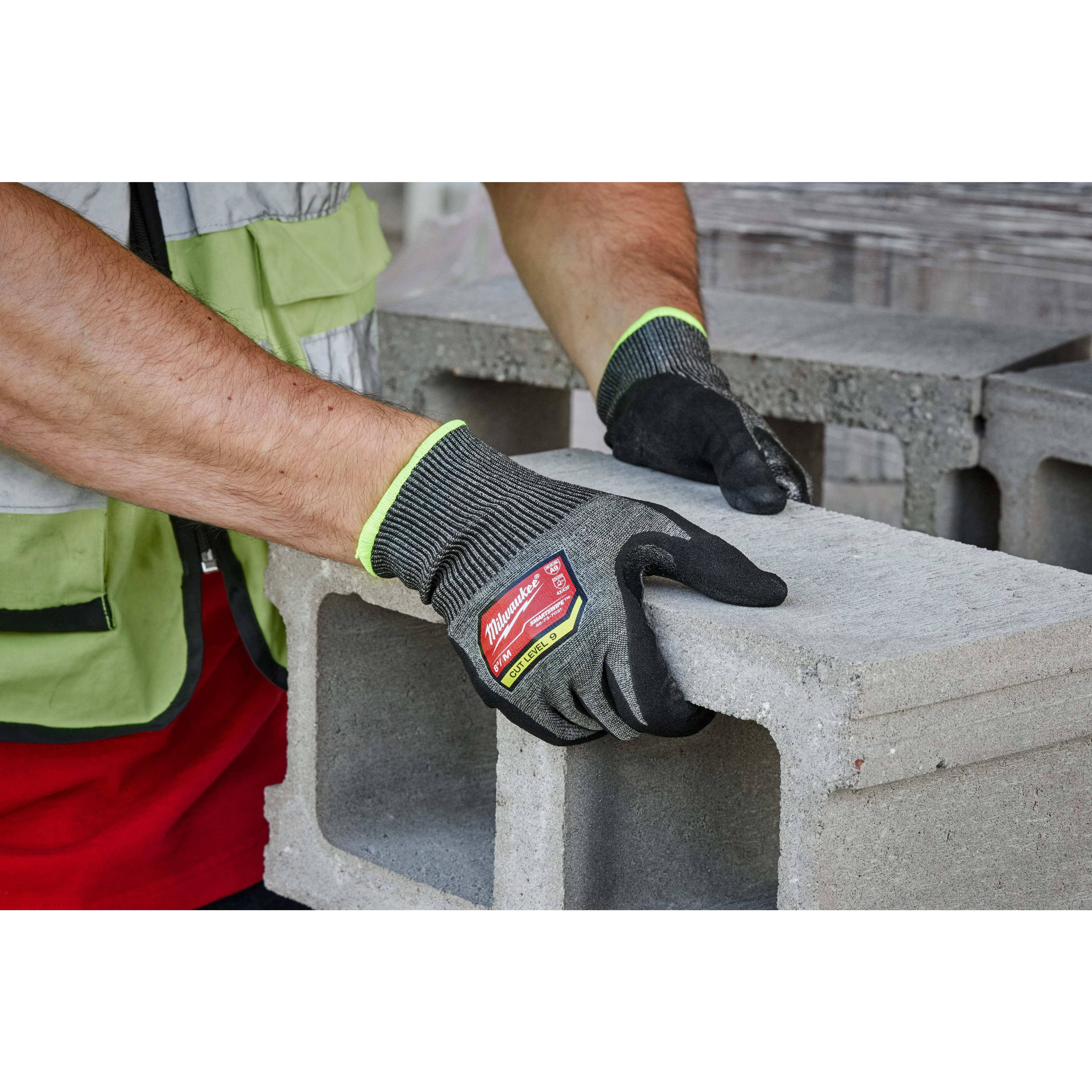 A person wears Cut Level 9 High-Dexterity Nitrile Dipped Gloves - M while handling a concrete cinder block. The gloves are black with green trim and a red label indicating their cut resistance level.