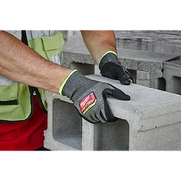 A person wears Cut Level 9 High-Dexterity Nitrile Dipped Gloves - M while handling a concrete cinder block. The gloves are black with green trim and a red label indicating their cut resistance level.